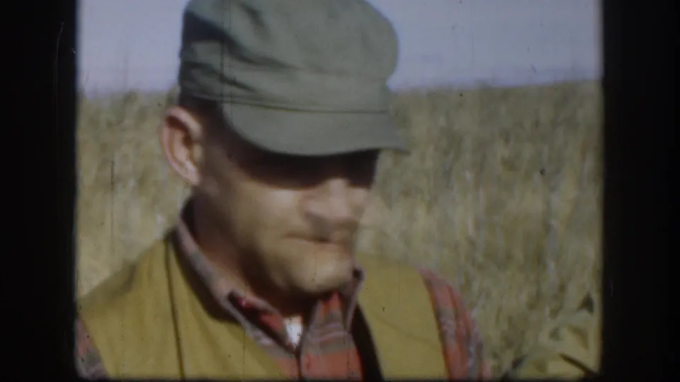 Close 8mm frame of a capped hunter in plaid and vest looking down in a dry Kansas field.