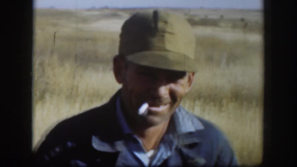 1958 Kansas home-movie frame of a capped adult hunter smoking in a dry open field.