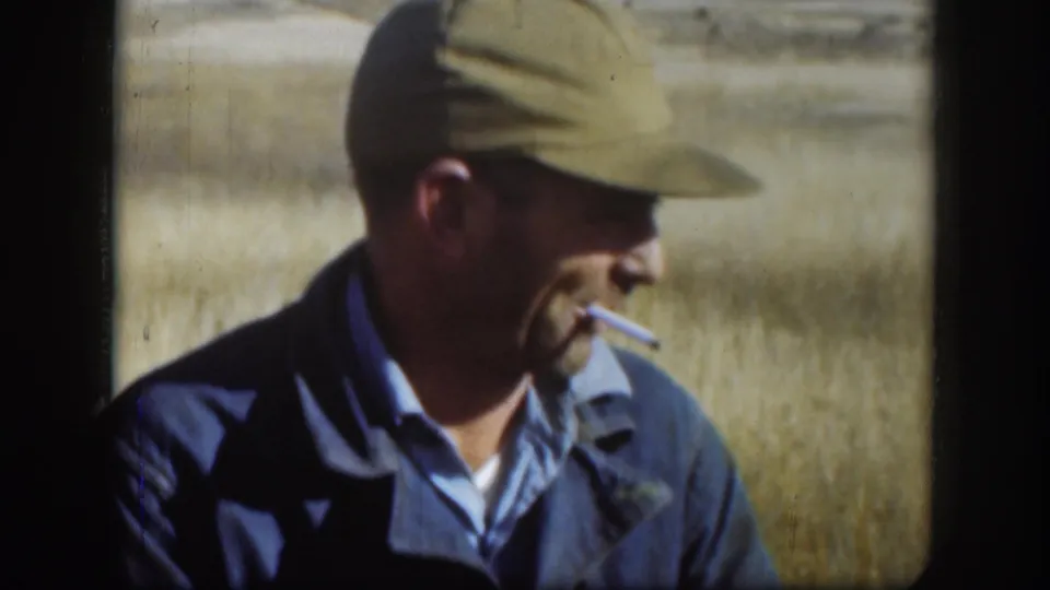 In Kansas 1958 home-movie footage, an adult man in a green cap and blue jacket smokes while standing in a dry field.