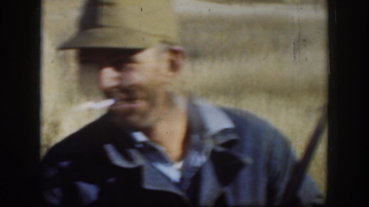 An adult man in a green cap and blue jacket appears slightly blurred while smiling with a cigarette in a Kansas field.