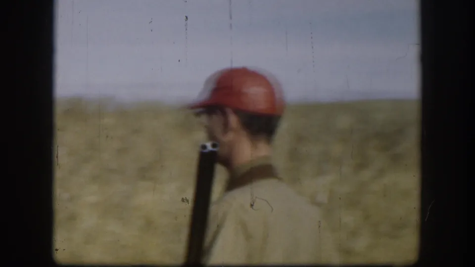 An adult man in a red cap stands in profile in a Kansas field with a long gun held upright near his shoulder.