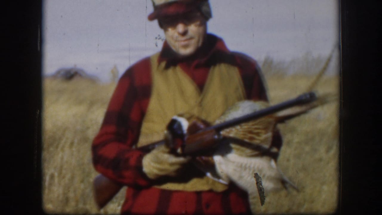 Man in plaid shirt and vest holds a rifle and harvested pheasant in a field.