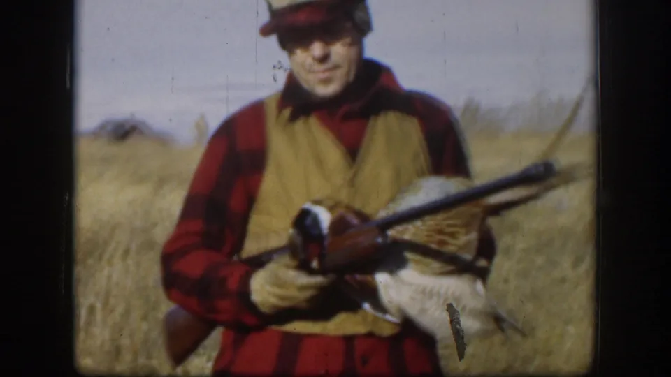 In the Kansas hunting scene, a man in plaid and a tan vest faces the camera holding a rifle and harvested pheasant.