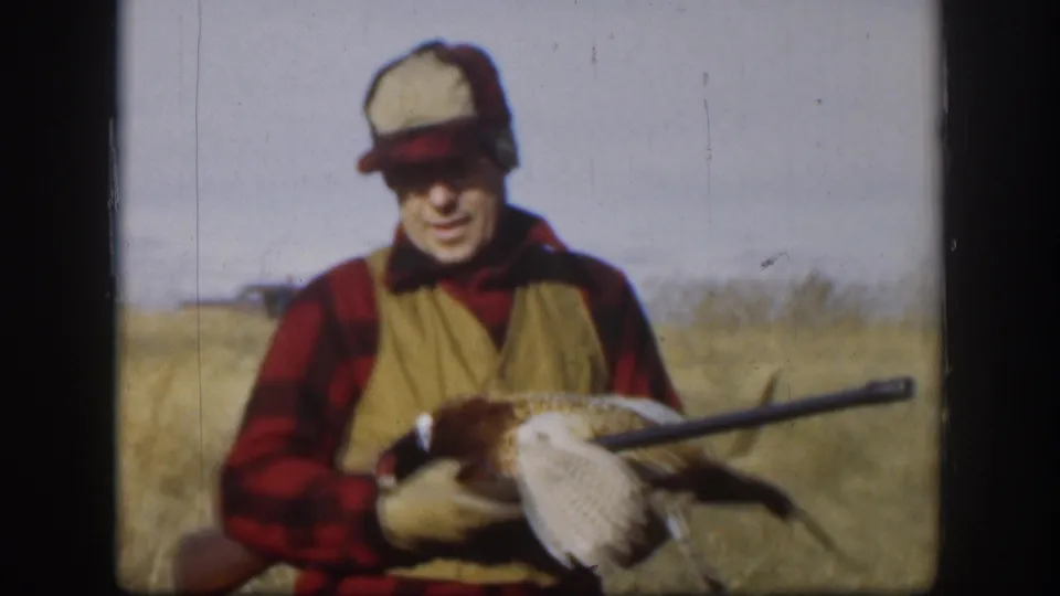 A smiling adult hunter in Kansas, 1958, poses in tall dry grass with a harvested pheasant draped near his rifle.