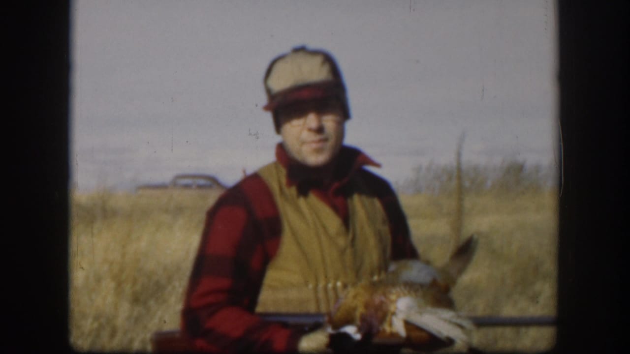 Hunter in plaid shirt and tan vest holds a pheasant and rifle in a dry field.