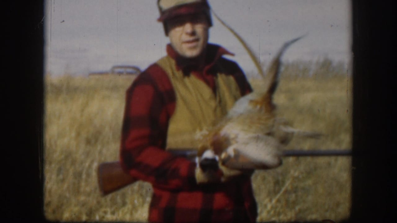Hunter holds up a pheasant while a rifle rests across his body in the field.