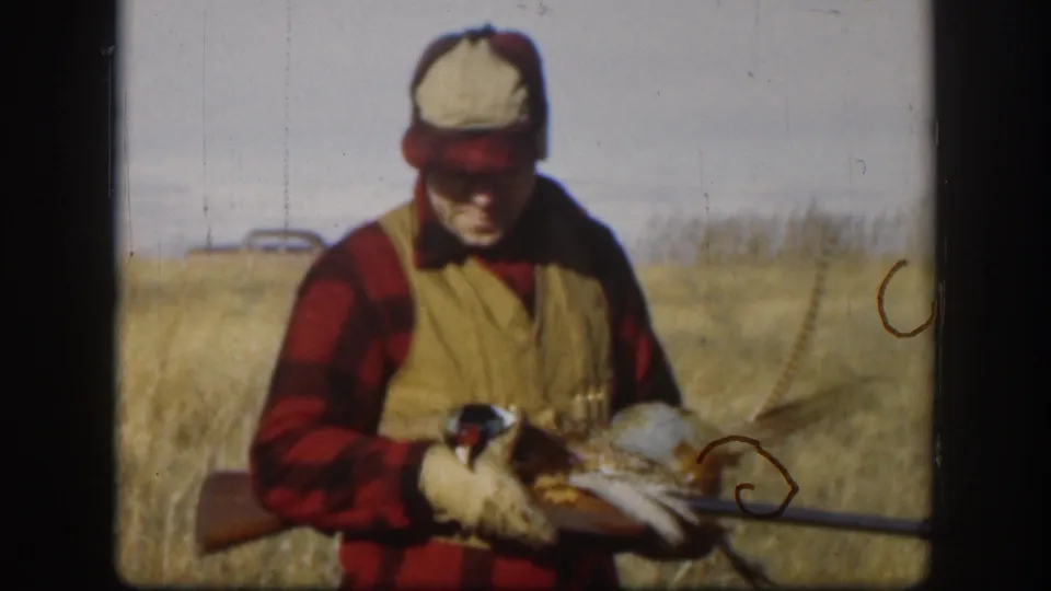 A hunter in cap and vest looks down at the pheasant he is holding, with his rifle visible across the lower frame.