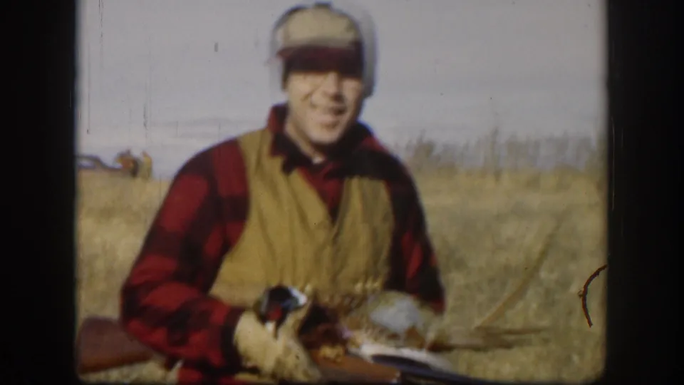 The hunter smiles toward the camera while holding a pheasant and rifle in a Kansas field, recorded on 8mm home-movie film.