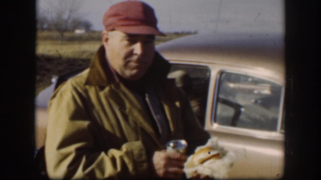Man in a red cap holding food and a can beside a rounded vintage car in a rural field.
