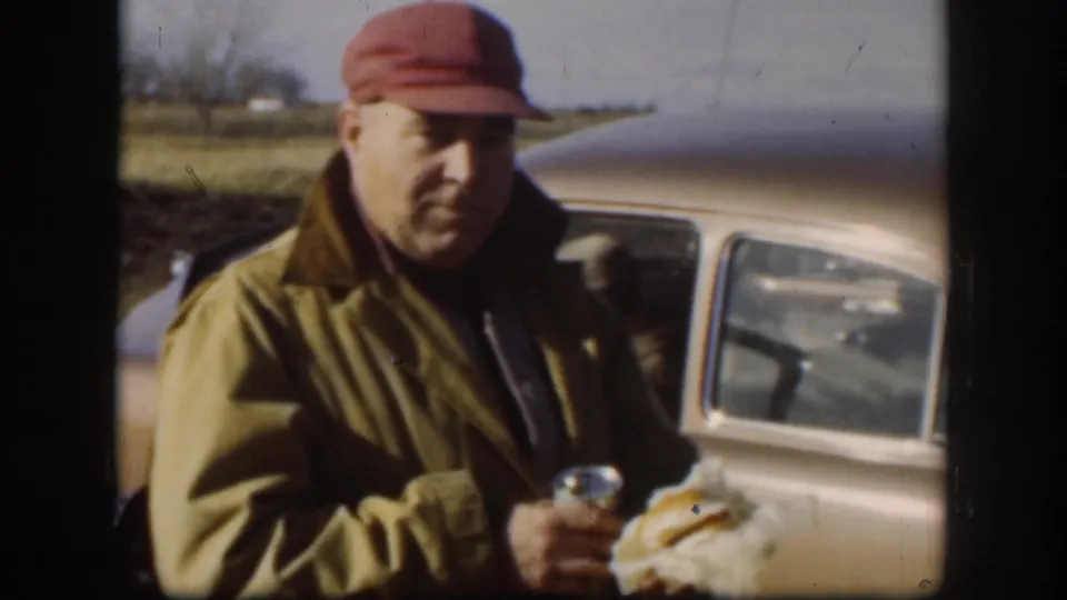 In supported 1958 Kansas home-movie footage, a man in a red cap and tan jacket stands beside a rounded vintage car with food and a can.