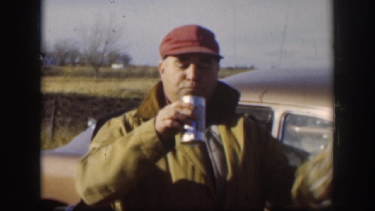 Man in a red cap drinks from a can while standing in front of a vintage car outdoors.