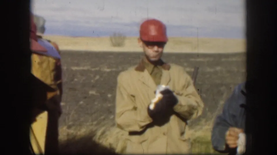 A man in a red cap and tan jacket holds a can in an open field, with other figures partly visible at the frame edges.