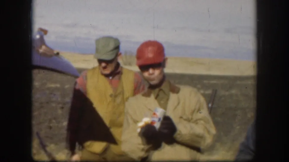Two adult men in caps stand together in a rural field, one holding a can, with part of a vintage car visible at the left edge.