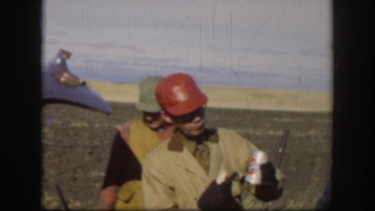 Two adult men outdoors, one in a red hard hat and sunglasses holding a can.