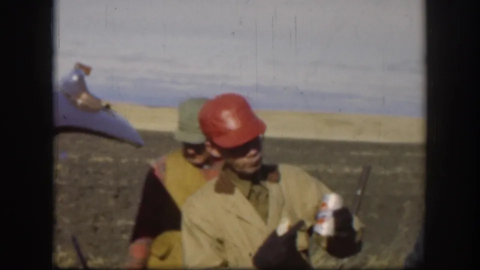 1958 Kansas home-movie frame of two adult men in a dusty outdoor setting, with one man in a red hard hat holding a canned beverage.