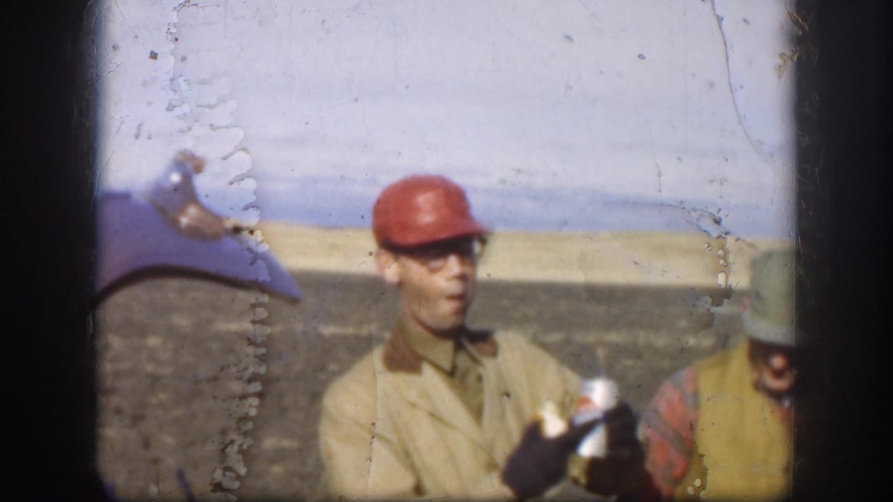Kansas 1958 home-movie frame showing an adult worker in a red hard hat and sunglasses holding a glass bottle in a rural outdoor setting.