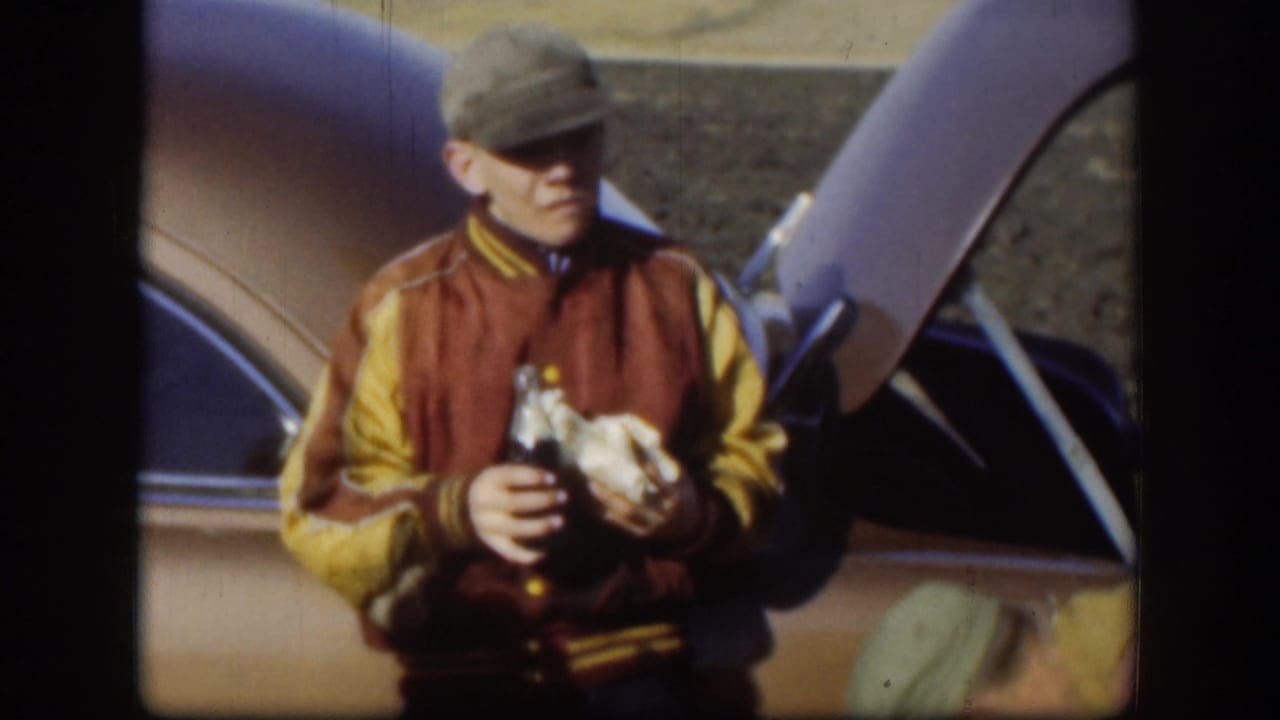 Kansas 1958 frame of an adult man in a cap and jacket holding a drink bottle and wrapped food beside a vehicle with its hood raised.