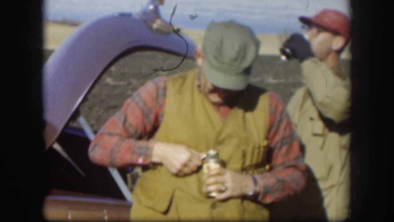Kansas 1958 home-movie frame showing two adult workers near an open vehicle hood, with one handling a glass bottle during a break.