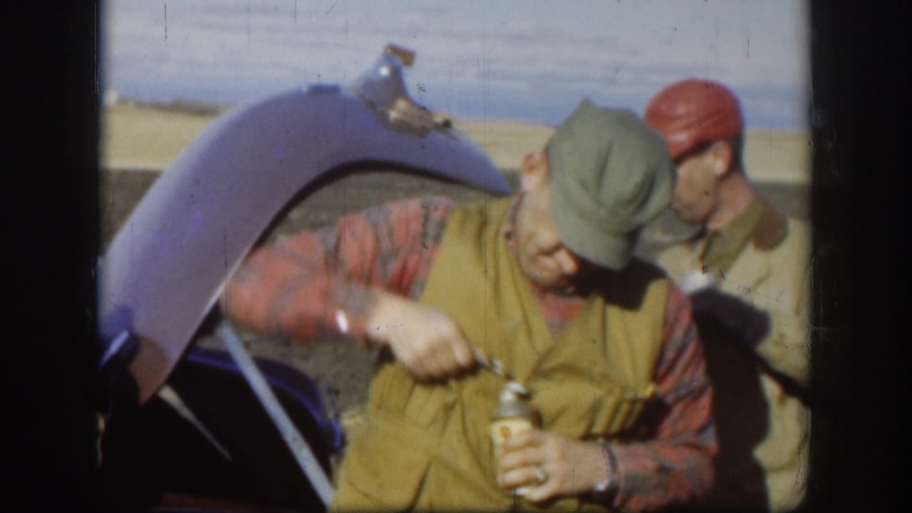 In rural Kansas 1958 home-movie footage, an adult hunter opens a drink near an open car trunk as another man in a red cap watches.