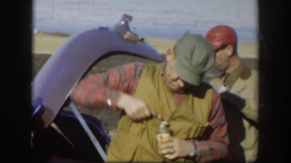 In rural Kansas 1958 home-movie footage, an adult hunter opens a drink near an open car trunk as another man in a red cap watches.