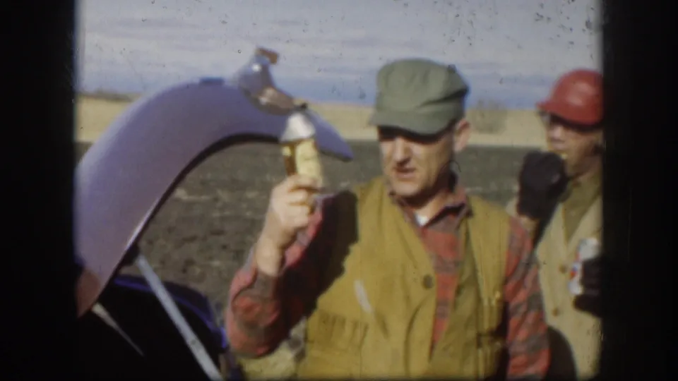 A Kansas hunting-trip frame from 1958 shows a man in a green cap holding a drink near an open trunk, with another hunter partly visible.
