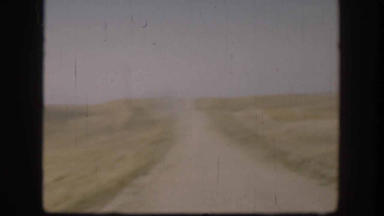 1958 Kansas 8mm frame showing a pale dirt road through open rural fields with soft focus and visible film scratches.