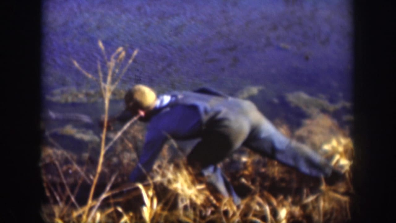 1958 Kansas home-movie frame of a man in a blue shirt and cap bending low while working in dry grass.