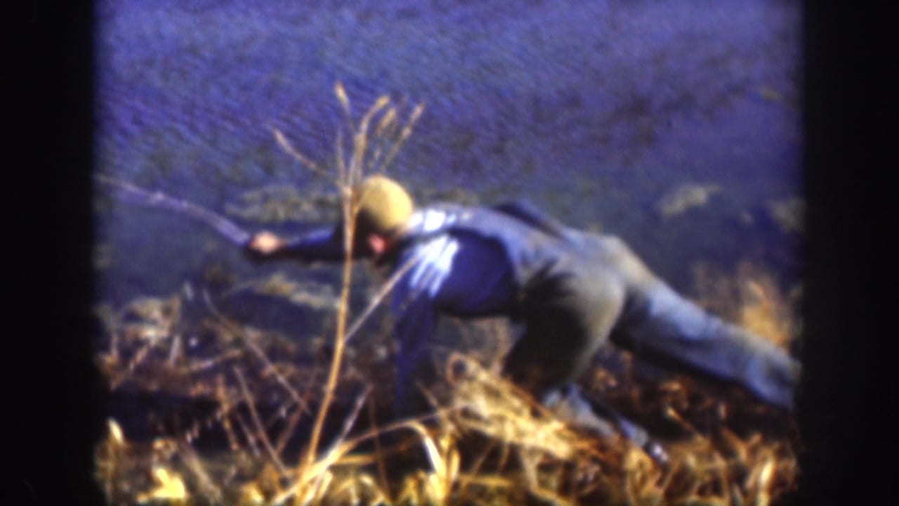 1958 Kansas home-movie frame showing a person in a blue shirt and light cap crouched low in shoreline grass beside water.