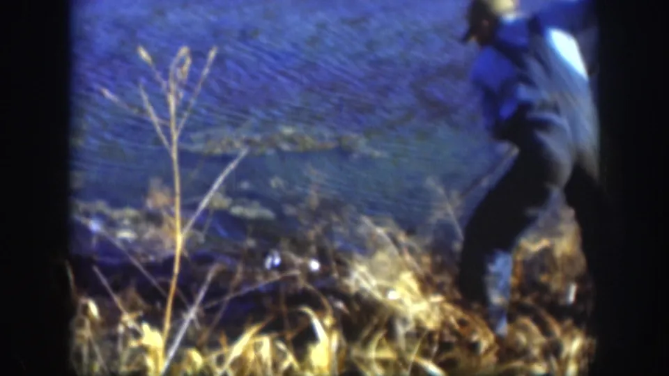 Kansas 1958 8mm frame showing a capped man partly at the lakeshore, moving beside reeds and water with a pole-like object nearby.