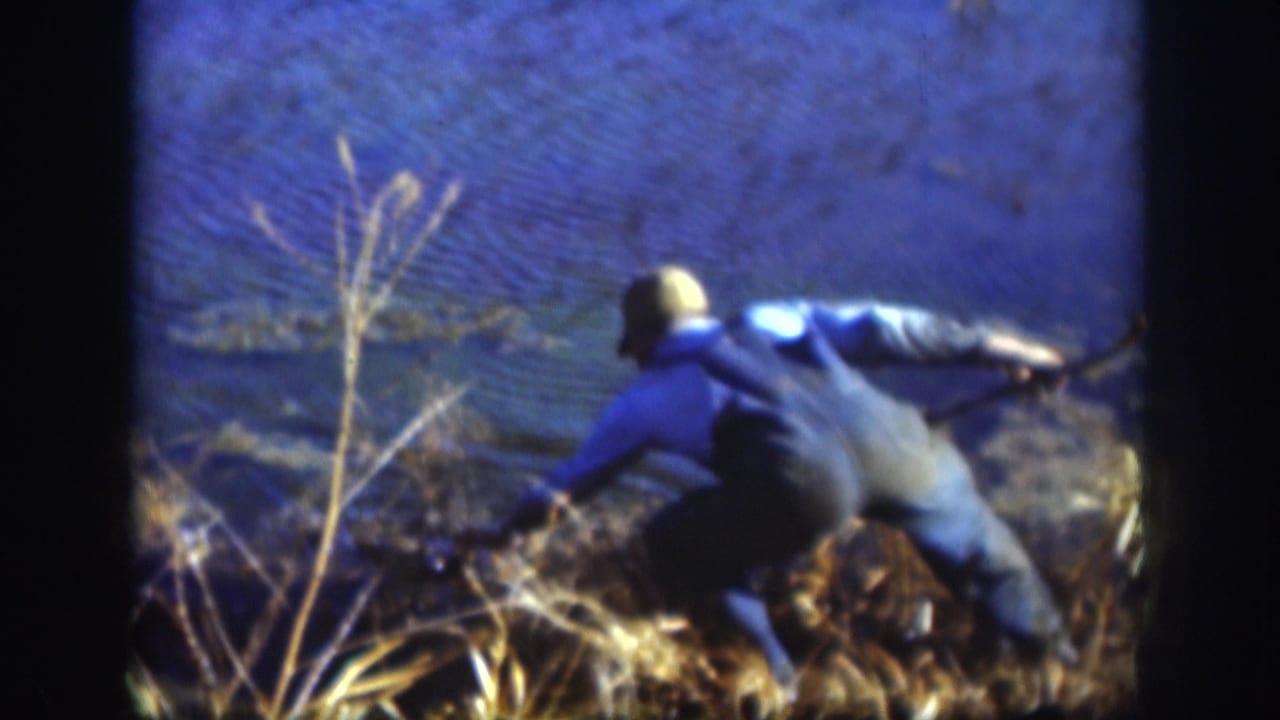 Kansas 1958 8mm frame showing a man in a blue shirt and cap leaning forward in tall reeds beside the lake.