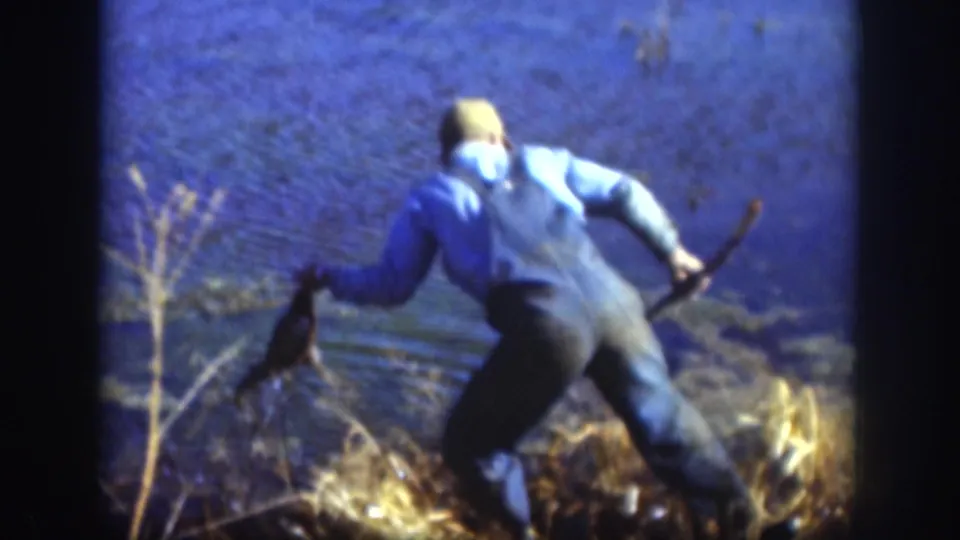 Kansas 1958 8mm frame showing a capped man at the lakeshore using a long pole to pull a duck from the water or reeds.