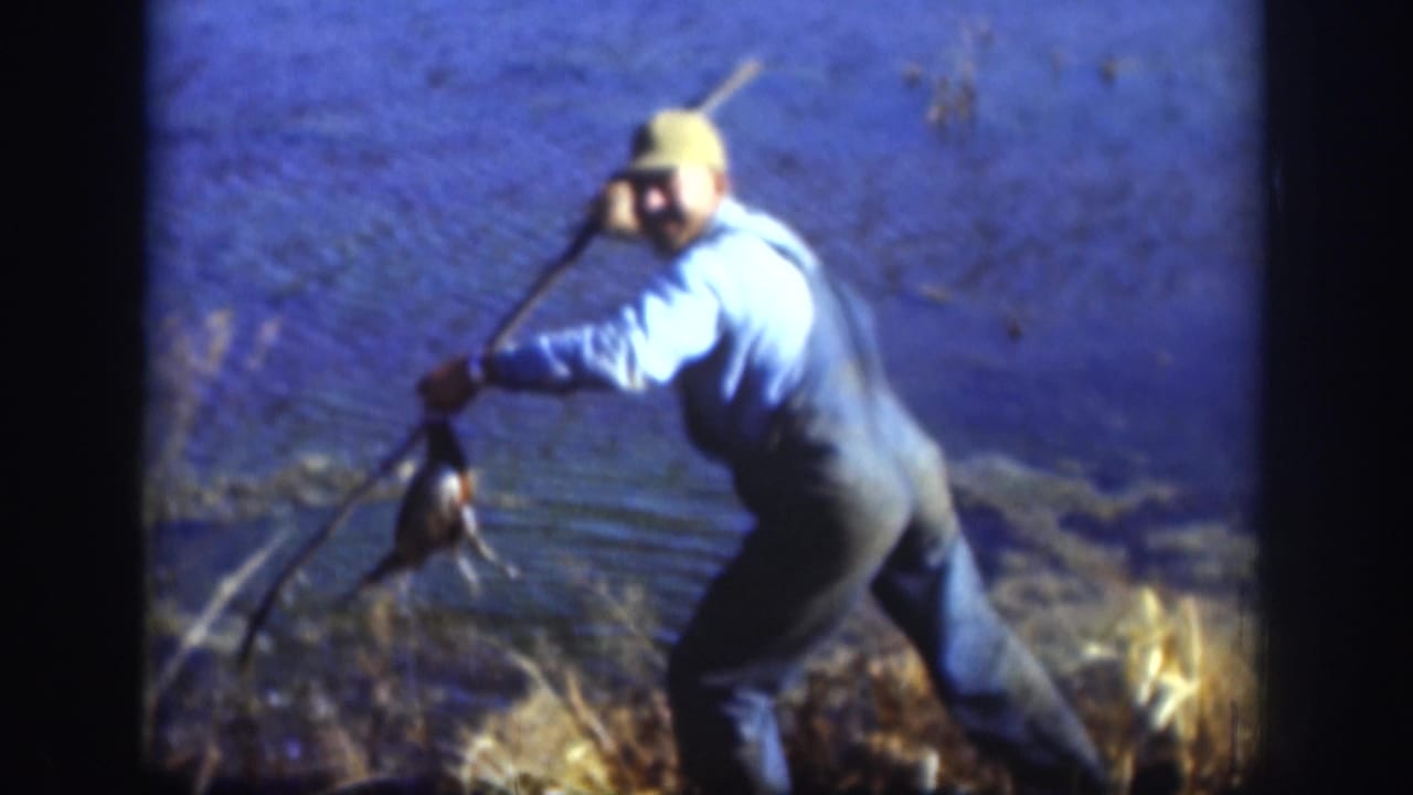 Man in overalls and cap holds a long gun and bird near marsh water.