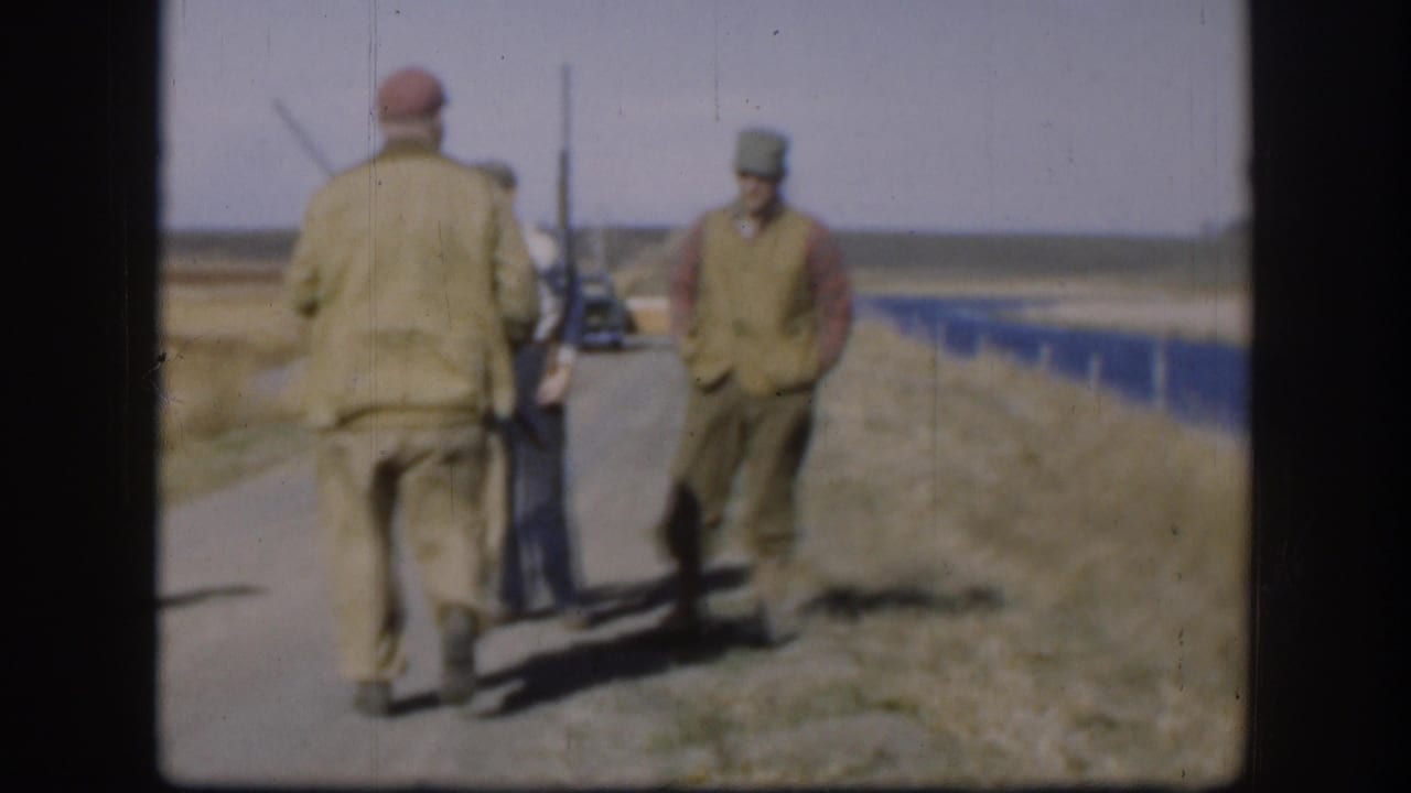 Two adult men in caps and outdoor clothing stand on a Kansas rural road, one carrying a long gun, with fields behind them.