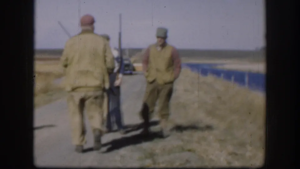 Two adult men in caps and outdoor clothing stand on a Kansas rural road, one carrying a long gun, with fields behind them.
