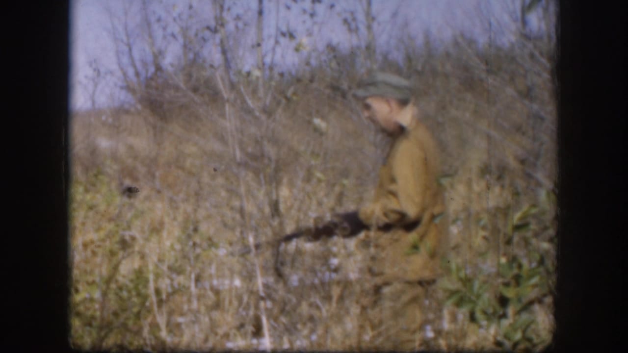 Adult hunter in a green cap and brown jacket moves through tall Kansas grass with a rifle in 1958 home-movie footage.