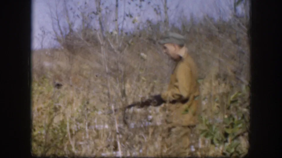 Adult hunter in a green cap and brown jacket moves through tall Kansas grass with a rifle in 1958 home-movie footage.