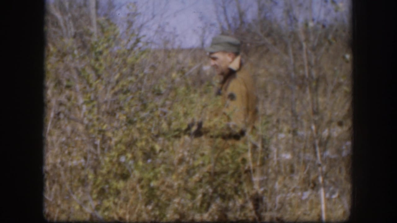Adult man in cap and brown jacket appears among brush and tall grass while holding a rifle in Kansas, 1958.