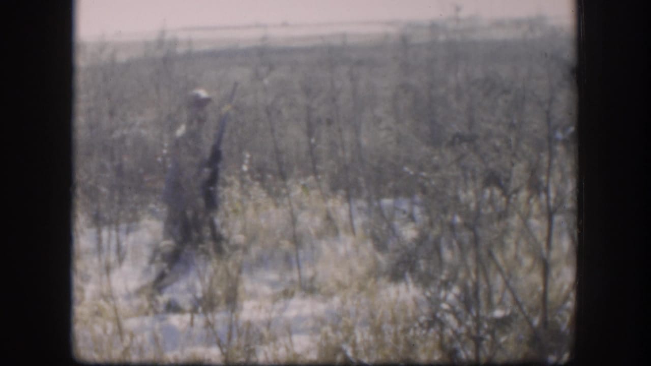 Adult hunter in rural Kansas appears in profile with a rifle while walking through pale winter grass and scattered snow.