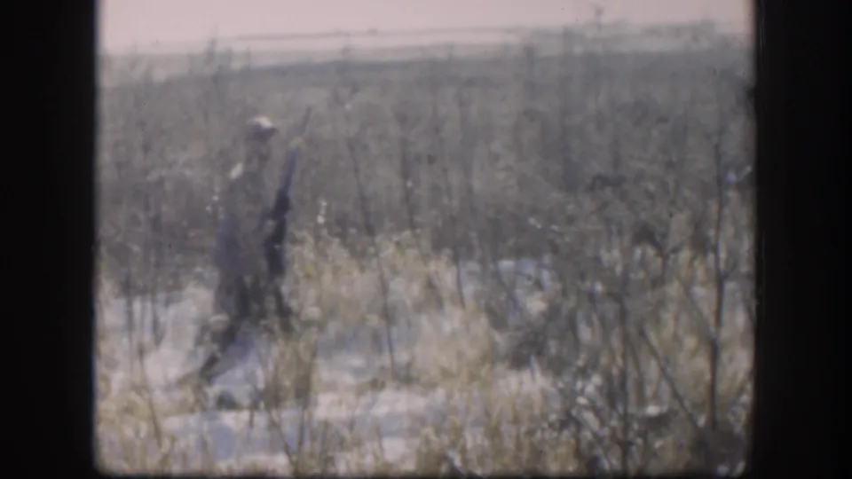Adult hunter in rural Kansas appears in profile with a rifle while walking through pale winter grass and scattered snow.