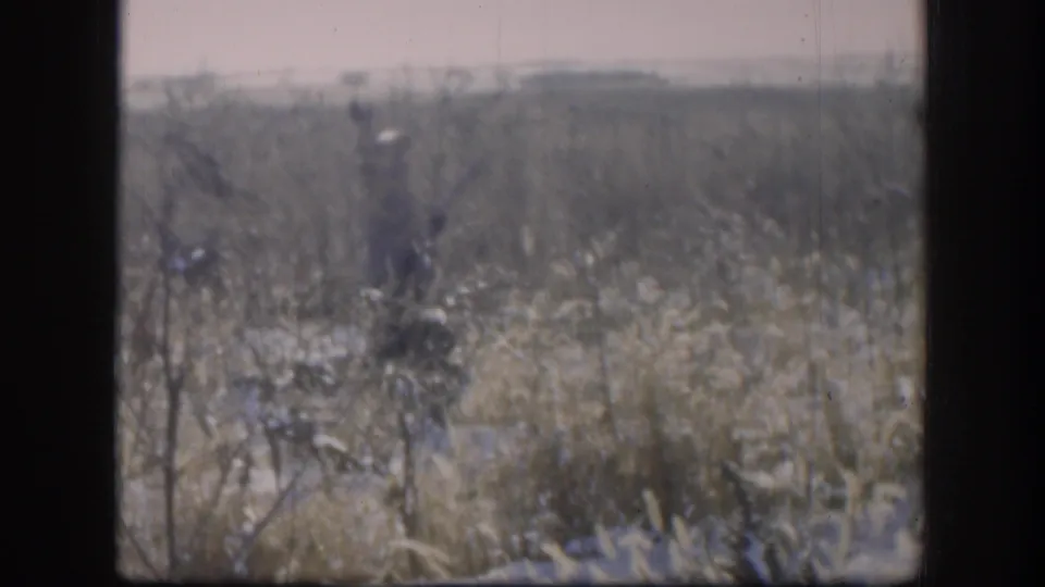 Adult hunter in 1958 Kansas stands among winter grass and snow patches, holding a rifle while facing the camera area.