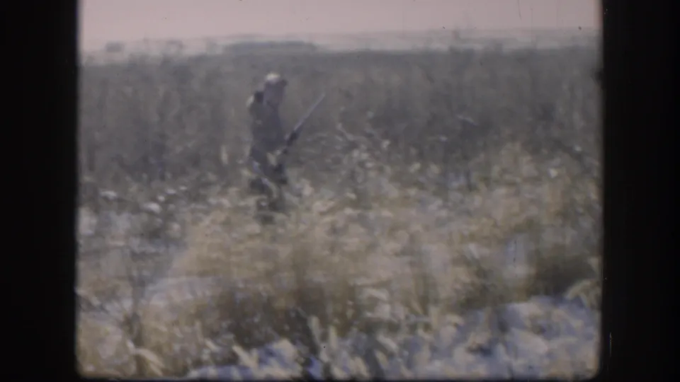 A hunter appears in a dry Kansas field in 1958 home-movie footage, carrying a shotgun across open grassland.