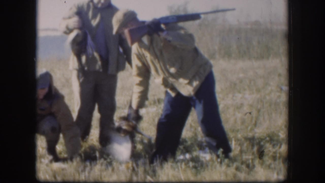Hunters in caps and field jackets gather in a dry Kansas field, with one person bending near a bird on the ground.