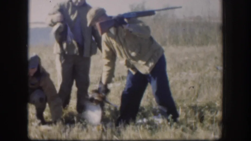 Hunters in caps and field jackets gather in a dry Kansas field, with one person bending near a bird on the ground.