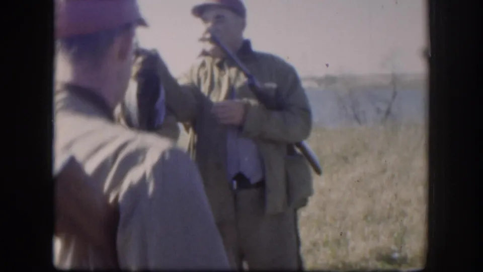 Two hunters converse in a dry Kansas field, with one holding game birds and a shotgun in 1958 home-movie footage.
