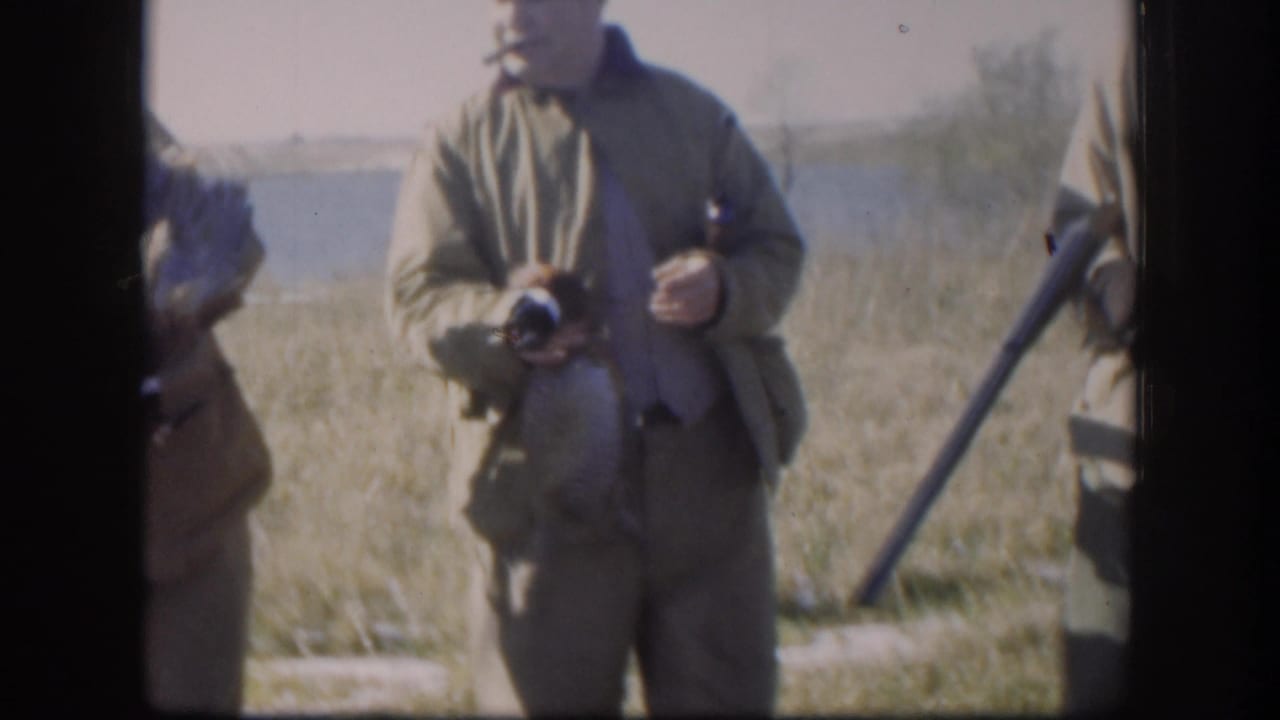 In 1958 Kansas home-movie footage, adult hunters in caps and jackets stand in a grassy field with shotguns and game birds near water.