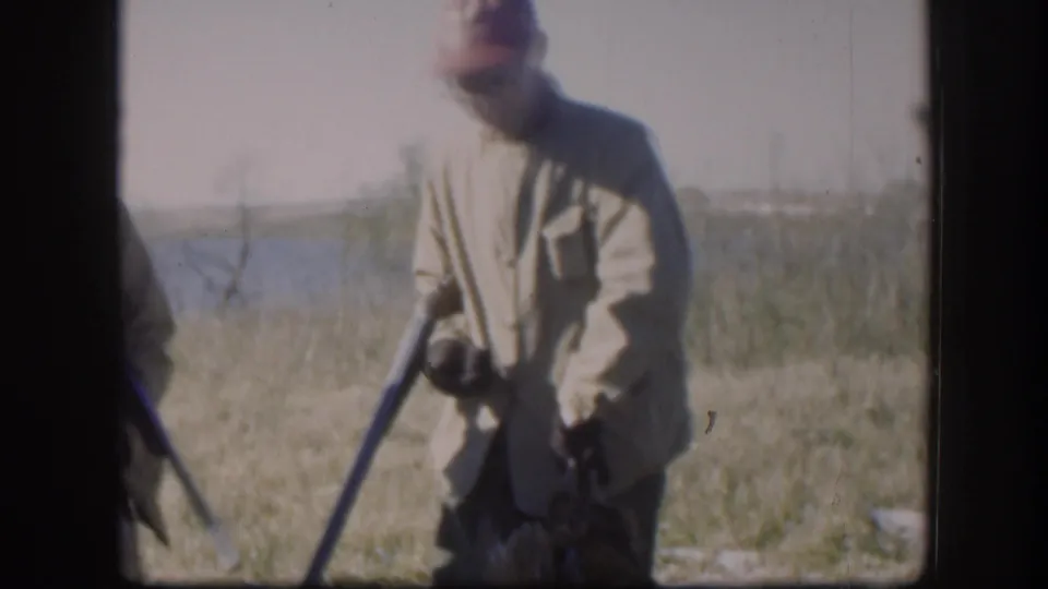 A Kansas hunter in mid-century field clothing stands near water, holding a shotgun and game bird for the 8mm camera.