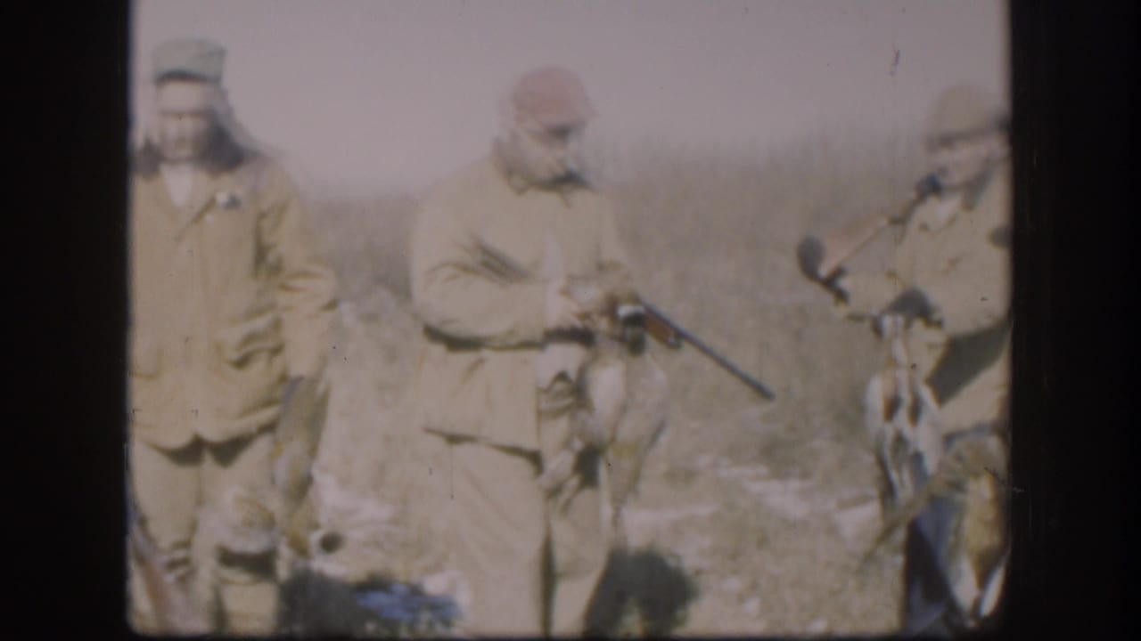 Adult men in field clothes stand with shotguns and game birds in a dry field.