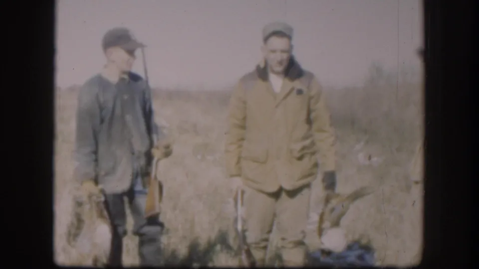 Two adult men in a Kansas field, one in a cap holding game birds and a shotgun, with another hunter beside him.