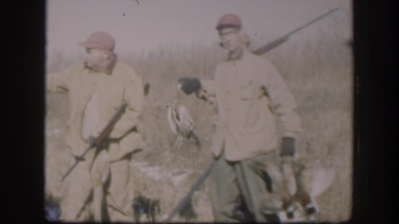 Two men in hunting clothes stand in a field with rifles and game birds.