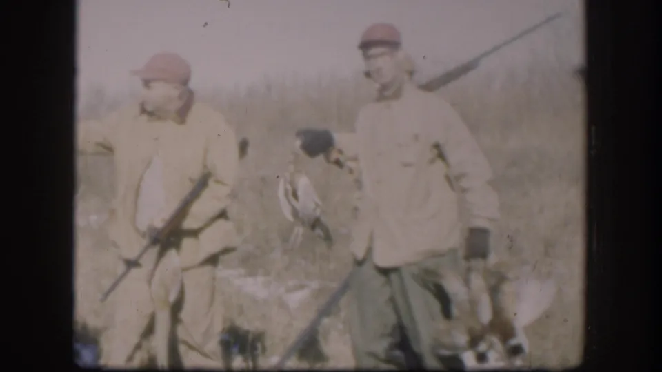 1958 Kansas home-movie frame of two men in red caps and tan jackets standing in a field with rifles and game birds.
